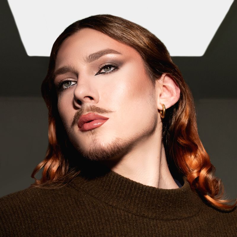 Striking headshot of an androgynous model with long hair, dramatic makeup, and stylish attire in a modern setting.